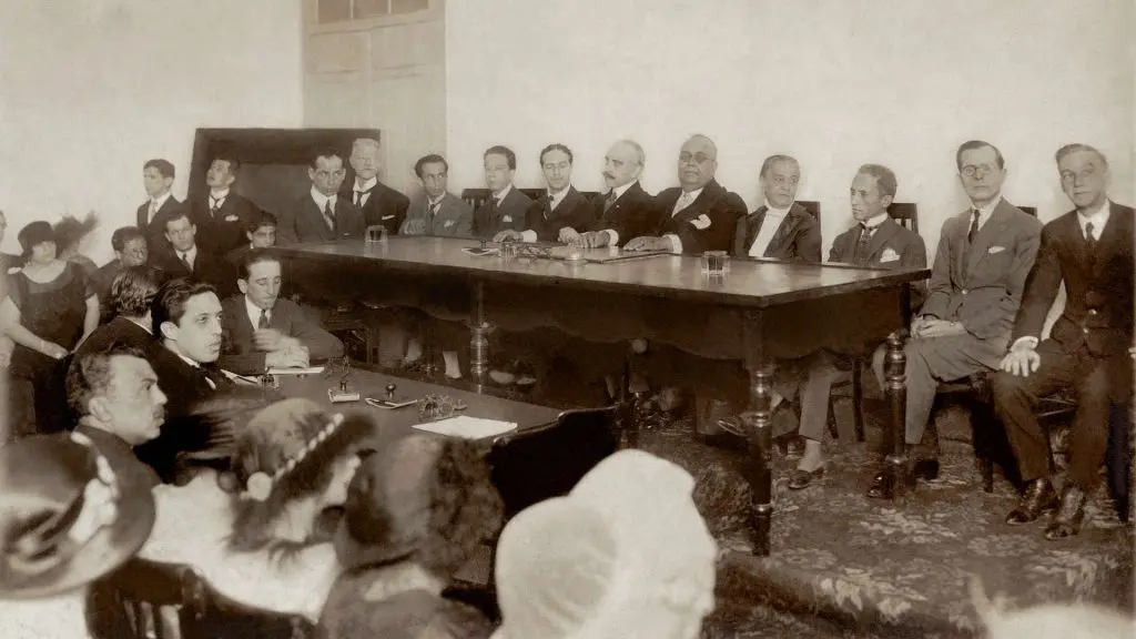 Reunião formal em ambiente interno, mesa de madeira, pessoas sentadas, discussão ou apresentação.