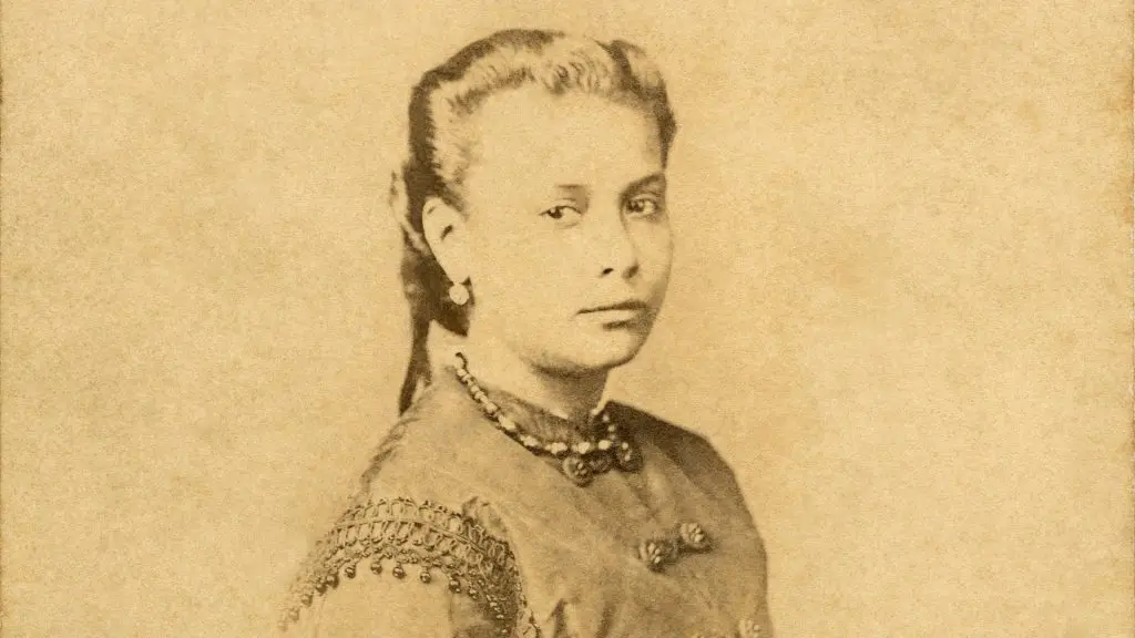 Mulher de perfil com vestido bordado e joia colgante, em tons de sepia.