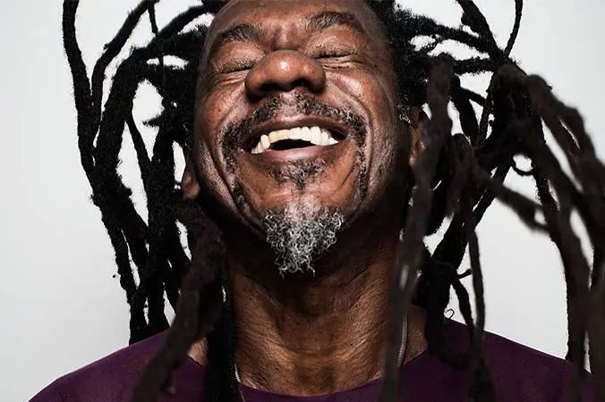 Pessoa sorridente com dreadlocks pretas e t-shirt roxa, fundo neutro.