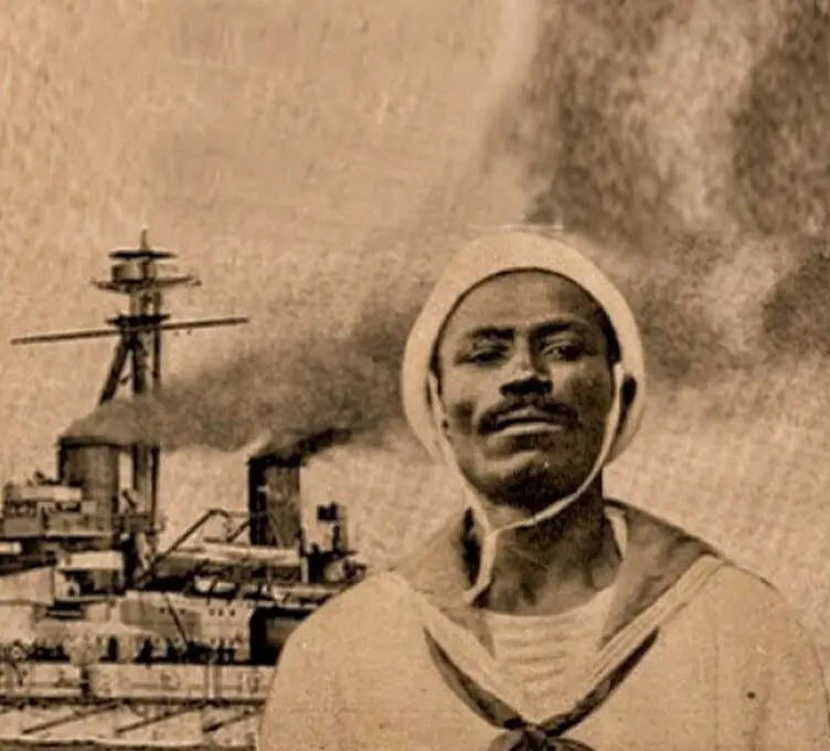 Marinheiro sorridente em uniforme de marinha, navio de guerra ao fundo em porto naval.