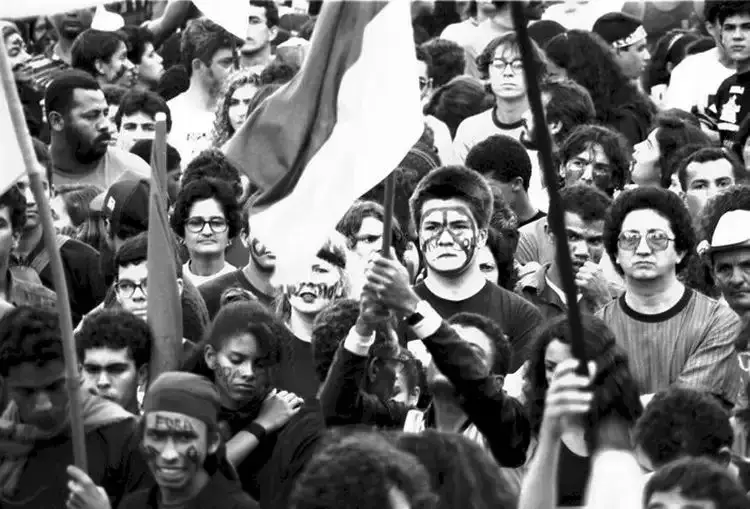 Multidão em protesto, rostos pintados, bandeiras e concentração.
