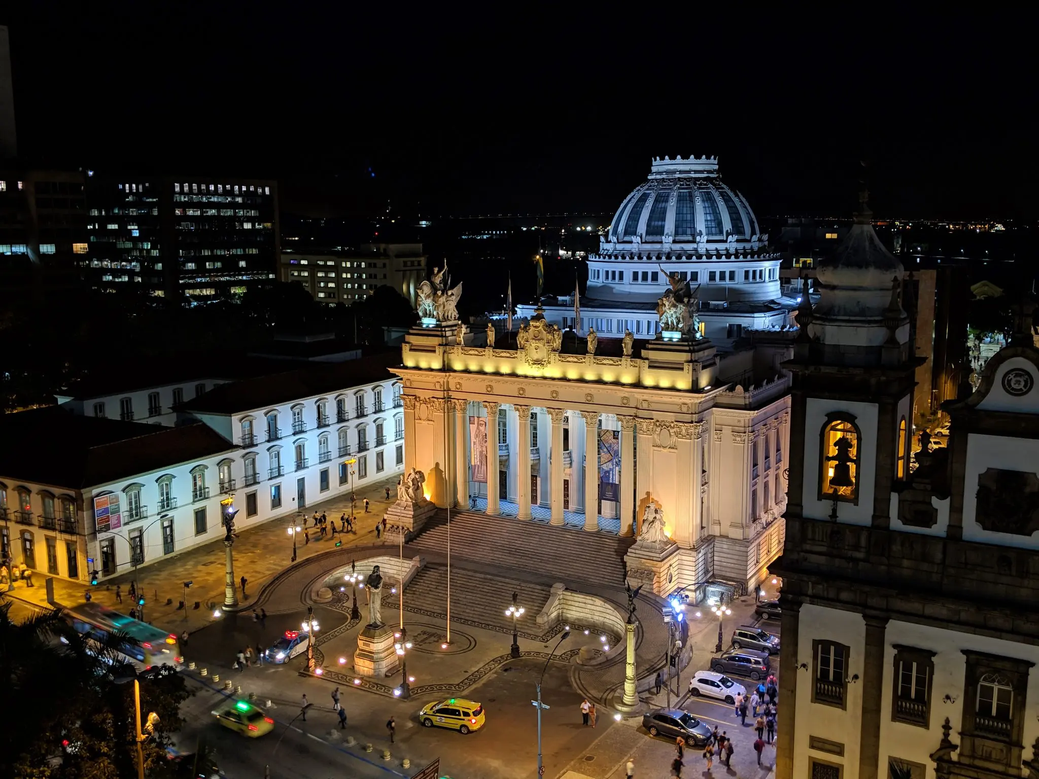 Palácio clássico iluminado à noite em praça urbana.