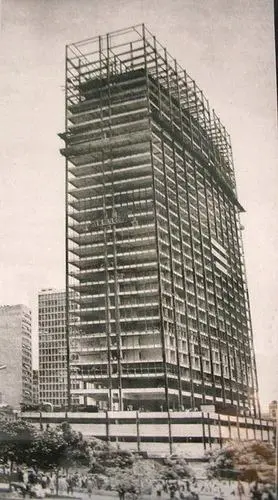 Edifício em construção com andaimes, prédios menores ao fundo.