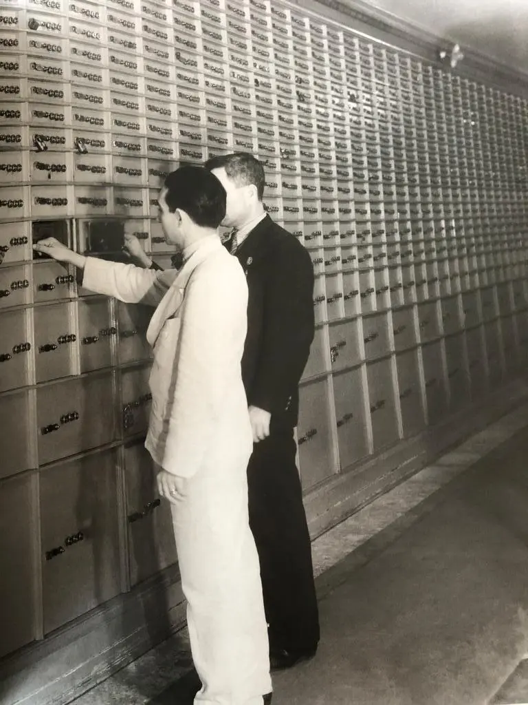 Dois homens em frente a gavetas metálicas, um em terno e outro em uniforme militar.