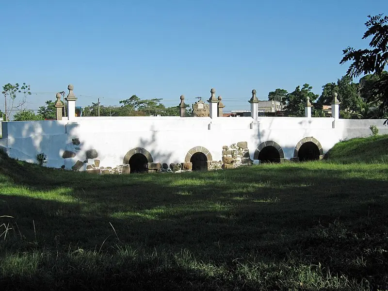 Ponte histórica em fazenda Santa Cruz, pedras e tijolos, arcos centrais, muro branco, céu azul claro.