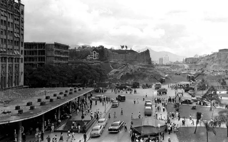 Cenário urbano com edifícios, pessoas caminhando, veículos e construção em construção.