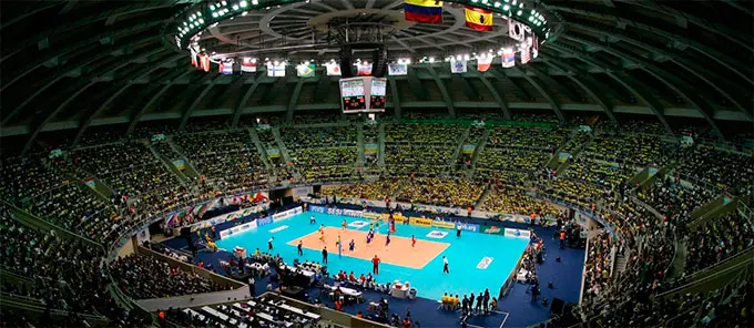 Estádio lotado durante jogo de vôlei, com torcedores em amarelo e bandeiras do Brasil penduradas.