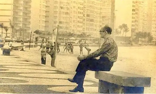 Homem em banco observa jogo de futebol na praia, com prédios altos ao fundo.