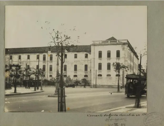 Convento antigo em preto e branco, com árvores e estrutura arquitetônica à esquerda, poste decorativo no centro, convento principal à direita, rua com veículos e pedestres na frente, demolido em 1911.