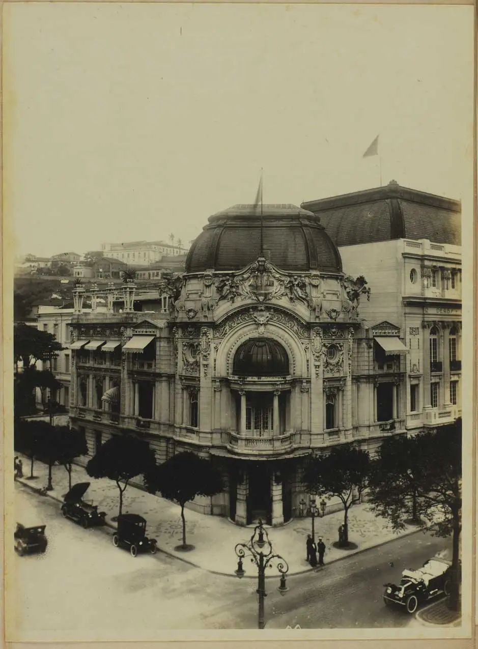 Edifício antigo em esquina com árvores e carros antigos.