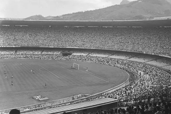 Estádio lotado durante jogo de futebol, com gramado verde, meta e público nas arquibancadas.