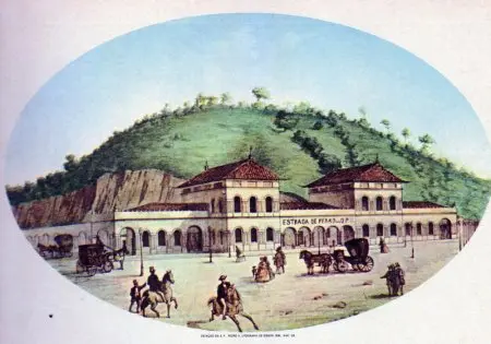 Edifícios de trens em frente a colina com carruagens e pessoas caminhando.