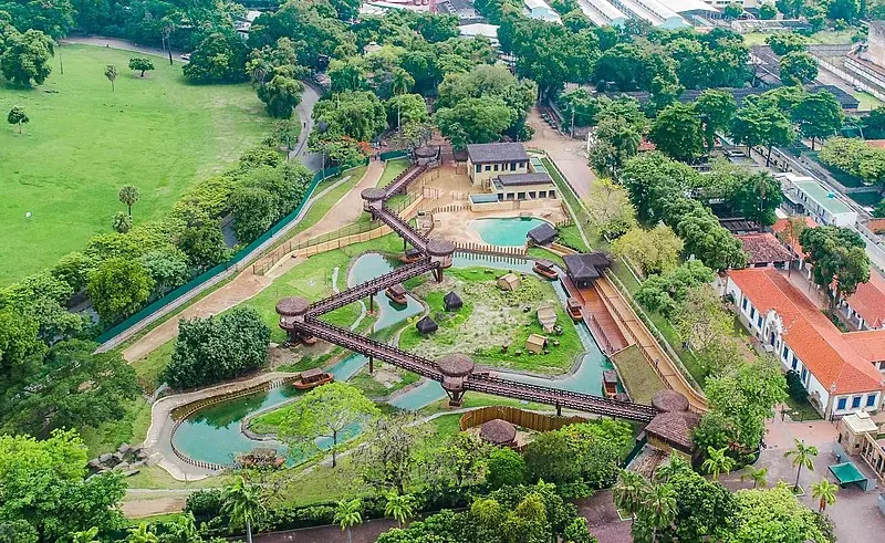 Parque zoológico com campos verdes, caminhos sobre lago e construções coloridas.