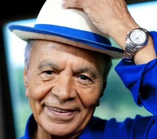 Homem sorridente com chapéu branco e relógio azul, fundo verde e azul desfocado.