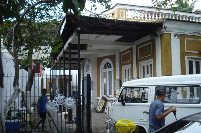 Casa colonial amarela com varanda coberta, árvores e van branca.