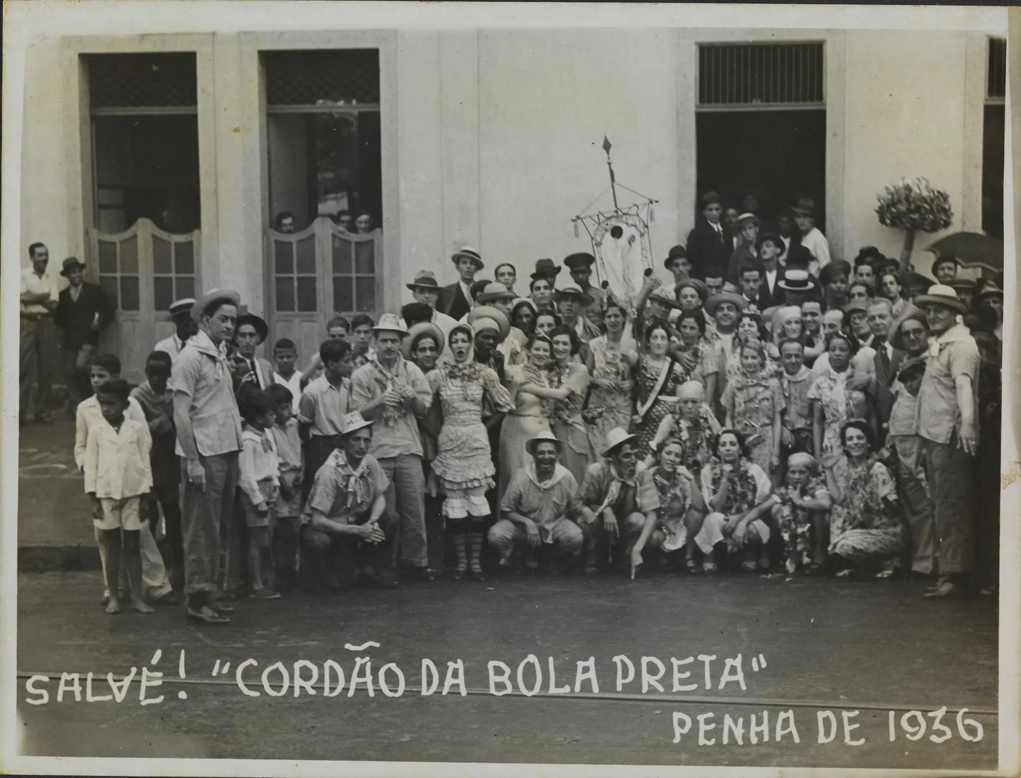 Grupo festivo em frente a edifício, com texto "SALVÉ! 'CORDÃO DA BOLA PRETA' PENHA DE 1936".
