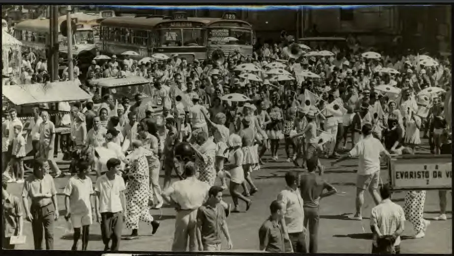 Multidão festiva em evento carnavalístico, com veículos e animação.
