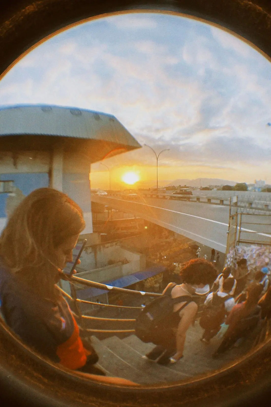 Pôr-do-sol com pessoa olhando para baixo, estrada e pedestres ao fundo.