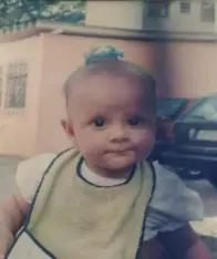 Criança sorridente em avental verde-amarelo, banco de madeira, carro preto, prédio com janelas grandes e grade metálica ao fundo.
