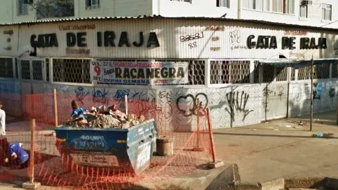 Prédio com fachada branca e grafites coloridos, placa "Cata de Irajá", contêiner azul e cerca vermelha.