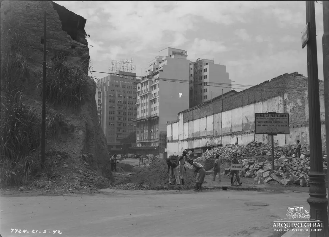 Trabalhadores em construção com penhasco e edifícios modernos ao fundo.