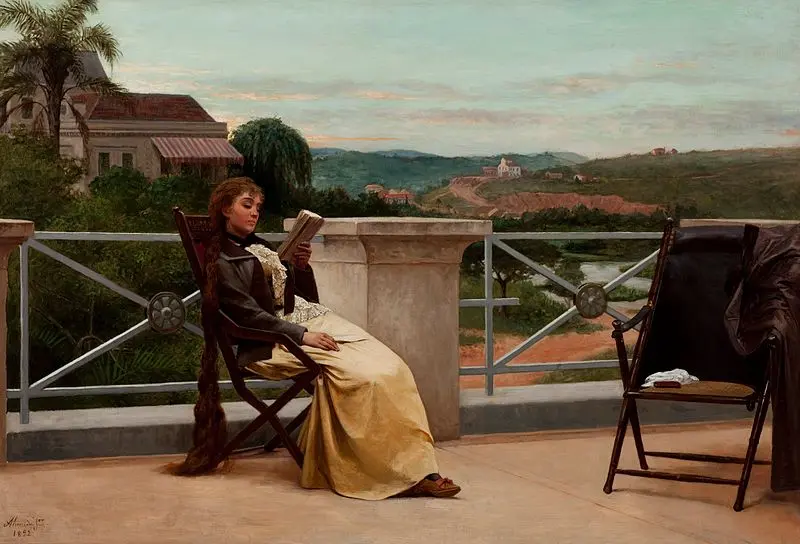 Mulher lendo livro em varanda com vista para paisagem rural.