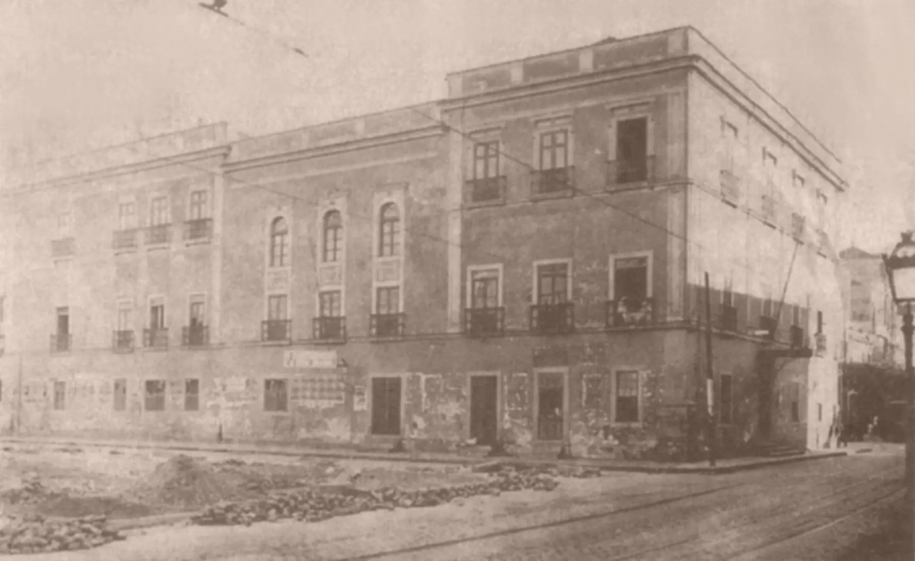 Edifício antigo com três andares, construções laterais e trilhos de bonde na frente.