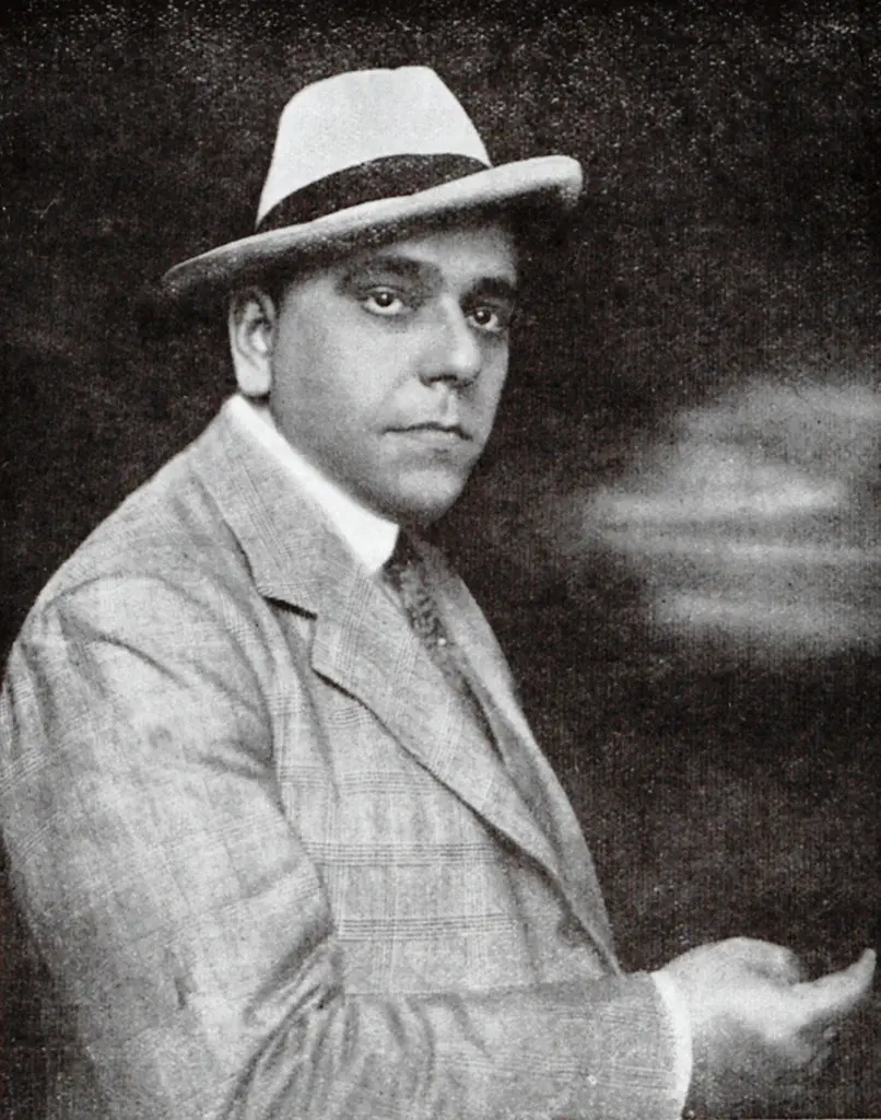 Homem com chapéu de palha e blazer xadrez, levantando a mão direita.