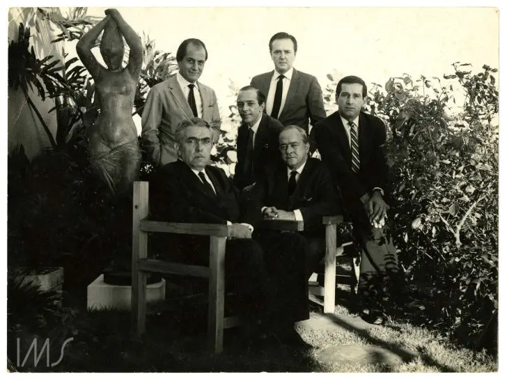 Homens em ternos em jardim com estátua humana à esquerda.