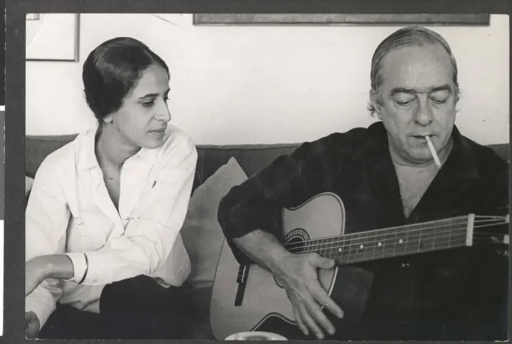 Dois amigos conversam em um sofá, um toca guitarra e fumaça.