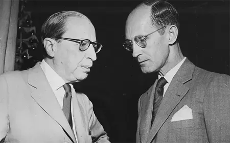 Duas pessoas em terno conversam em ambiente formal.