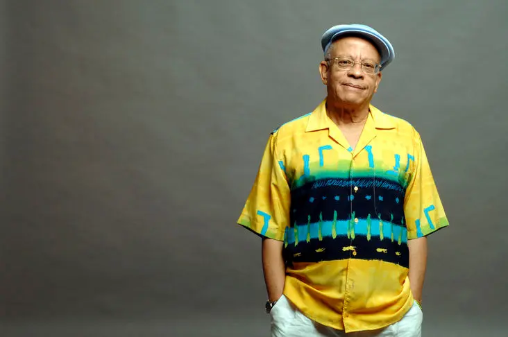 Homem sorridente em camisa amarela com padrões azuis e pretos, chapéu azul claro e óculos escuros, fundo cinza neutro.