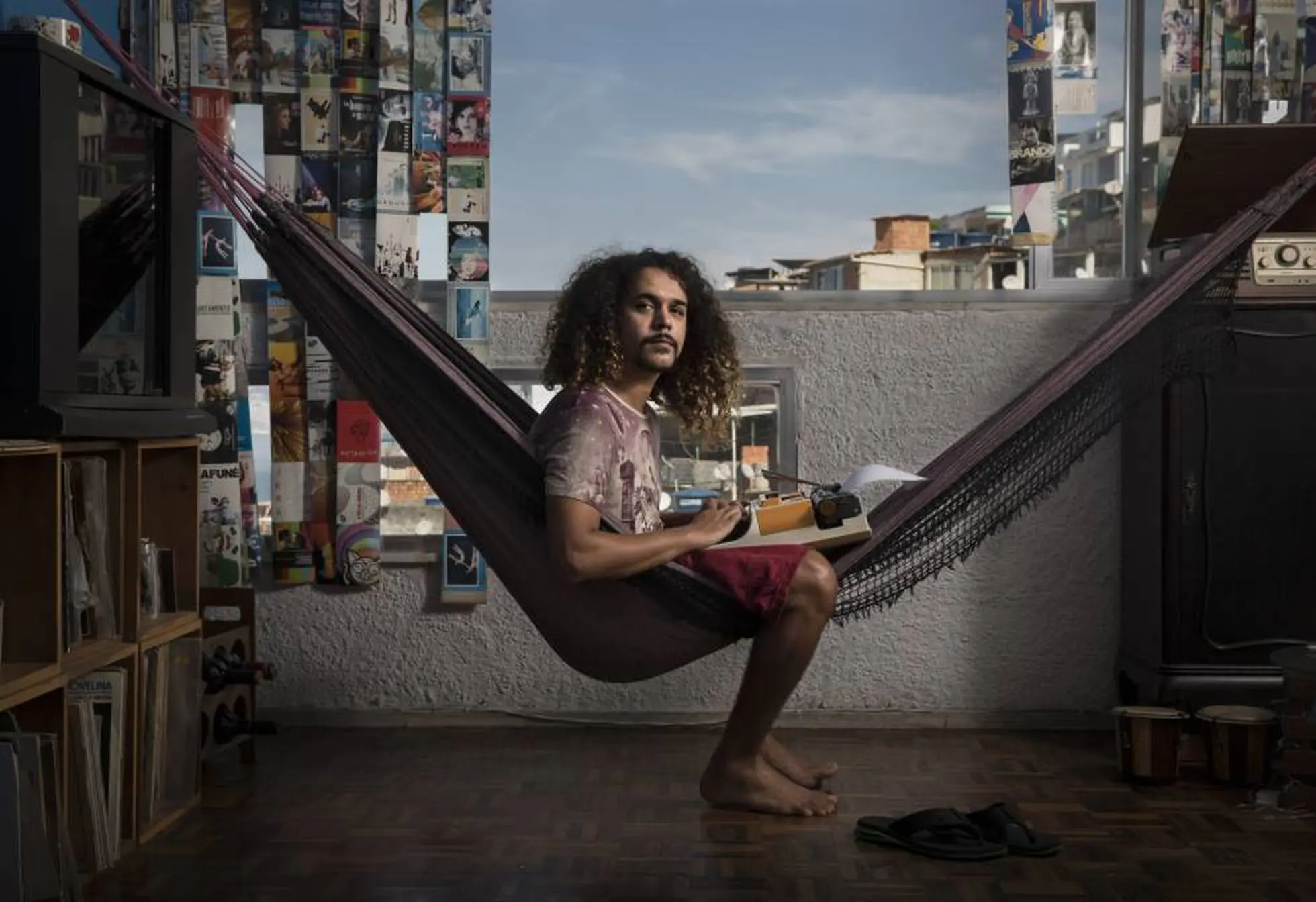 Homem em hammock com teclado de máquina de escrever, livros e sandálias ao redor.