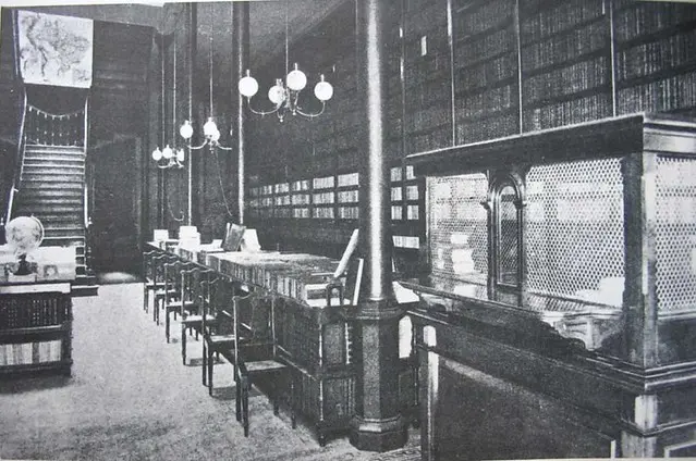 Biblioteca antiga com escada, livros empilhados, mesas e luminárias.