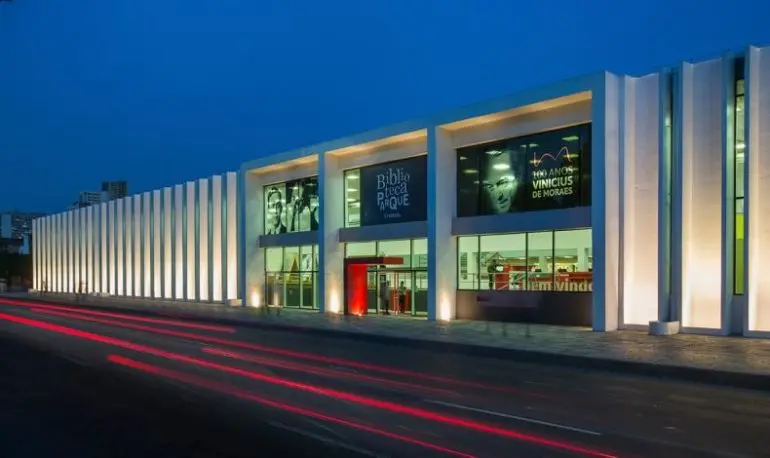 Edifício moderno à noite com fachada iluminada e entrada marcada como "Biblioteca Parque".