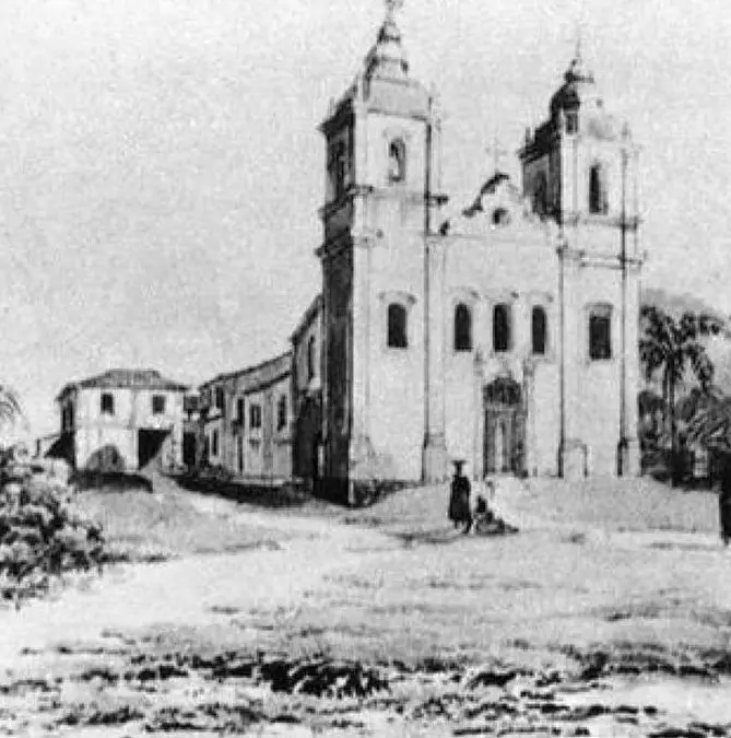 Igreja histórica com torre e cúpula, edifícios laterais e pessoas observando.