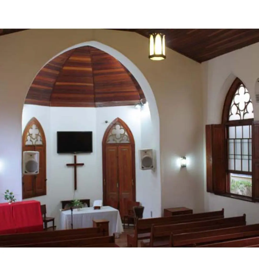 Interior de igreja com mesa vermelha, crucifixo, porta fechada, alto-falantes, televisão, vitrais e madeira.