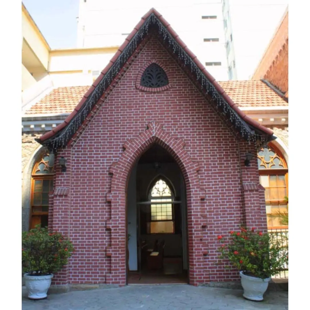 Capela de tijolos vermelhos com telhado de telhas vermelhas, janelas de vidro colorido e vasos de plantas ornamentais.