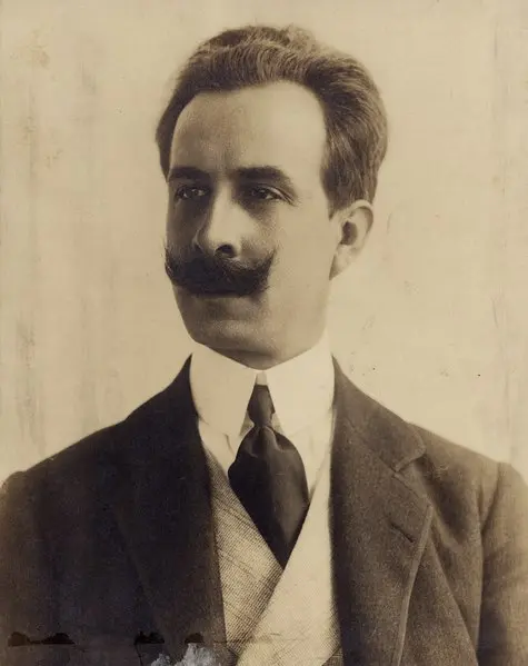 Homem elegante em terno clássico, barba bem cuidada, fundo neutro.
