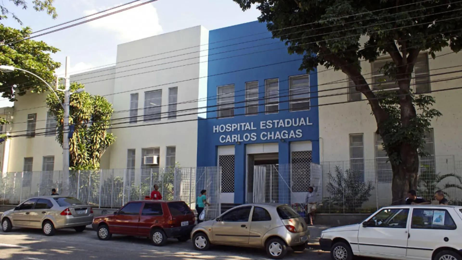 Hospital Estadual Carlos Chagas com prédios branco e azul, carros estacionados e árvores ao redor.