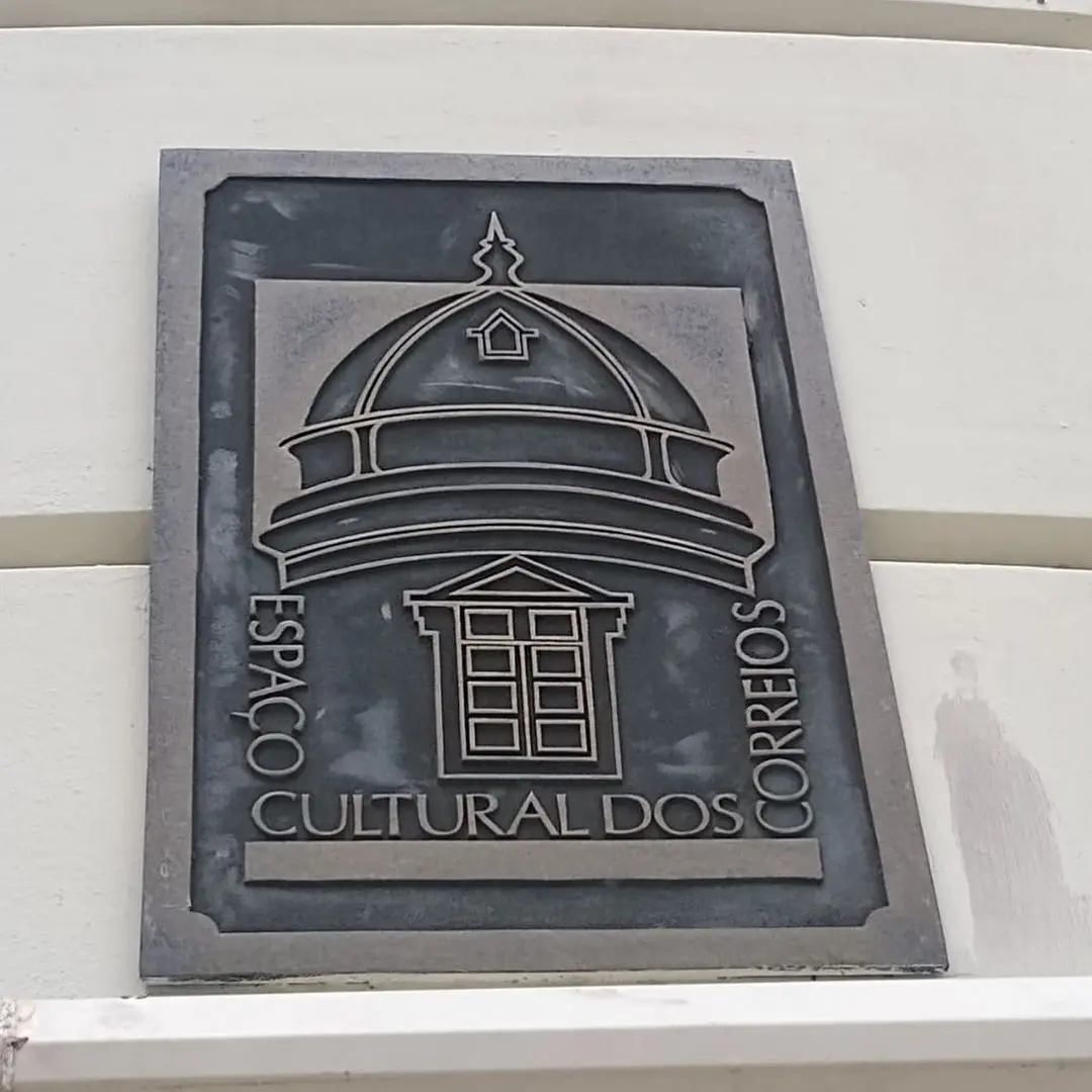 Placa metálica com desenho de construção cultural dos Correios.