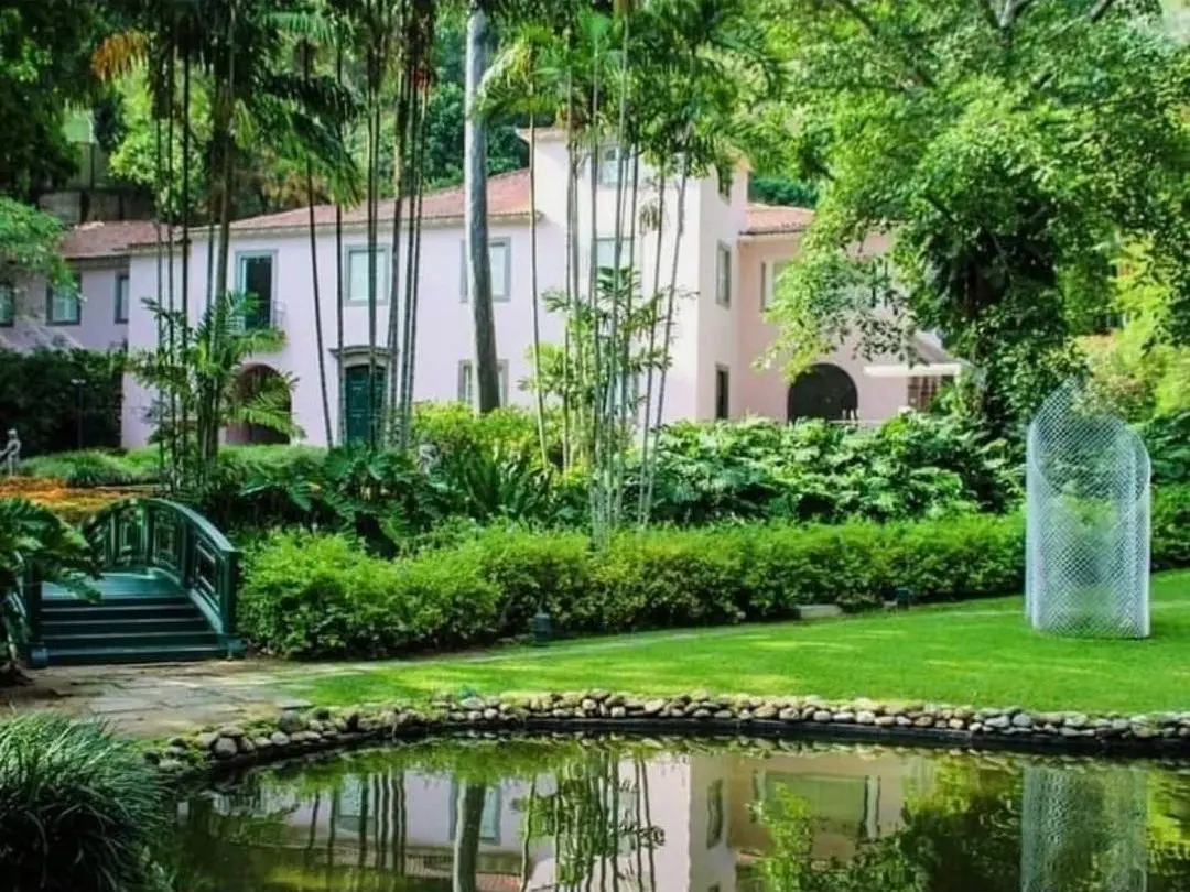 Jardim com casa, ponte verde, lago e estrutura transparente.