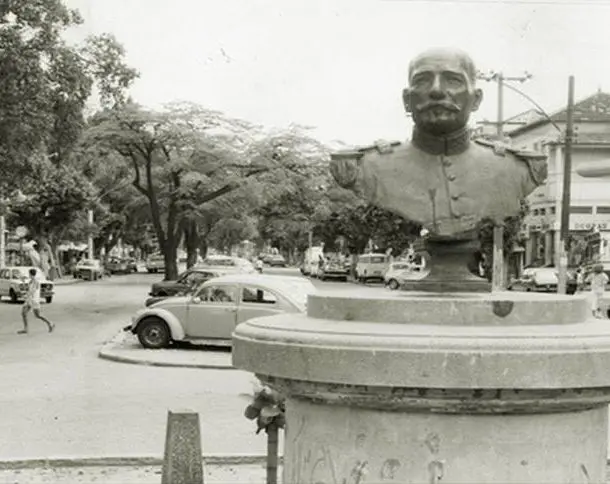 Busto militar em praça movimentada, com árvores e carros antigos.
