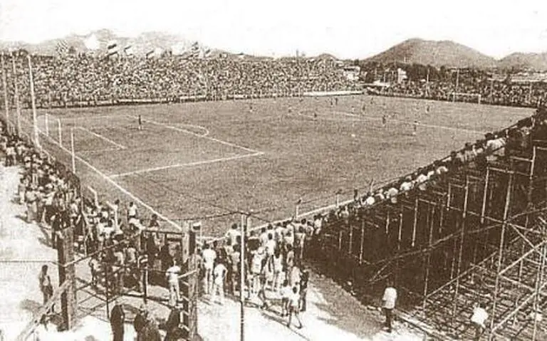 Estádio de futebol antigo com torcida e montanhas ao fundo.