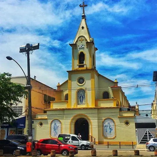 Igreja amarela em cenário urbano com árvore e prédios ao fundo.