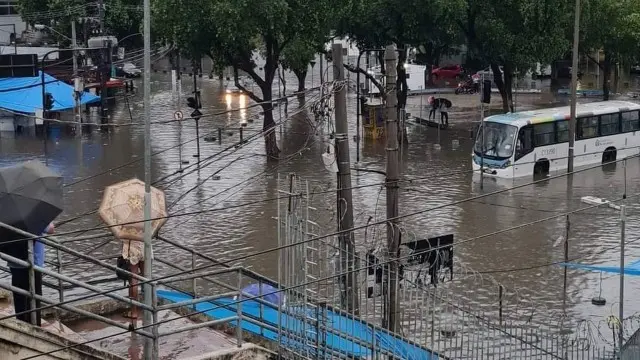Rua inundada em Marechal Hermes, com homem segurando guarda-chuva azul e ônibus parado na água.