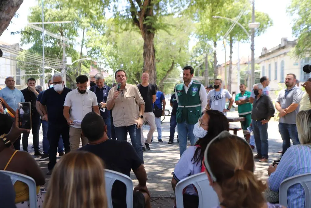 Pessoas sentadas em cadeiras brancas em um evento ao ar livre, com árvore e edifícios ao fundo.