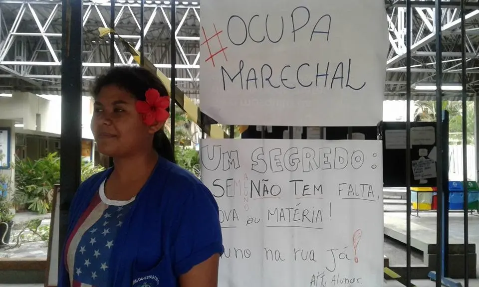 Mulher em frente a portão metálico com panfletos sobre ocupação e segredo.