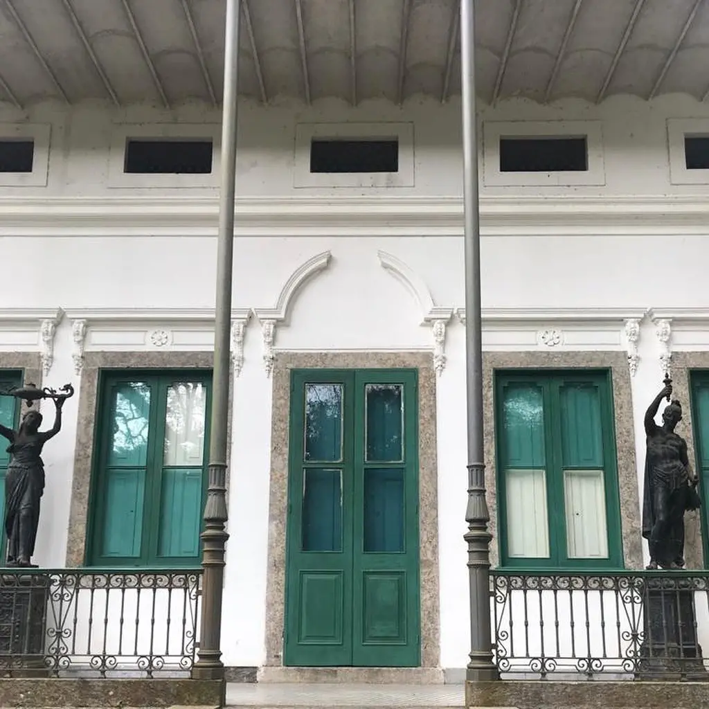 Edifício histórico com estátuas femininas e masculina, grades decorativas e janelas verdes.