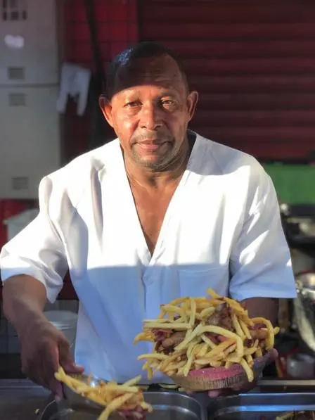 Homem sorrindo segurando hambúrguer e batatas fritas, camisa branca aberta, parede vermelha e janela com grades metálicas ao fundo.